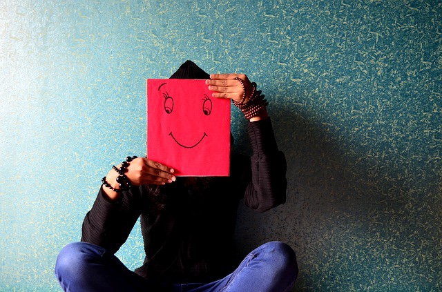 African-American person is seated and holding a red piece of paper over his or her face with a smiley face drawn on it.
