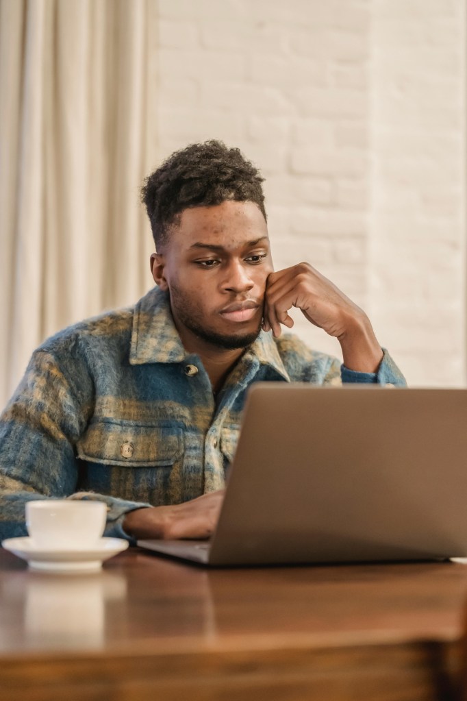 An African American man is seated at a table with a coffee mug and laptop. He is looking at the laptop and appears to be thinking. 