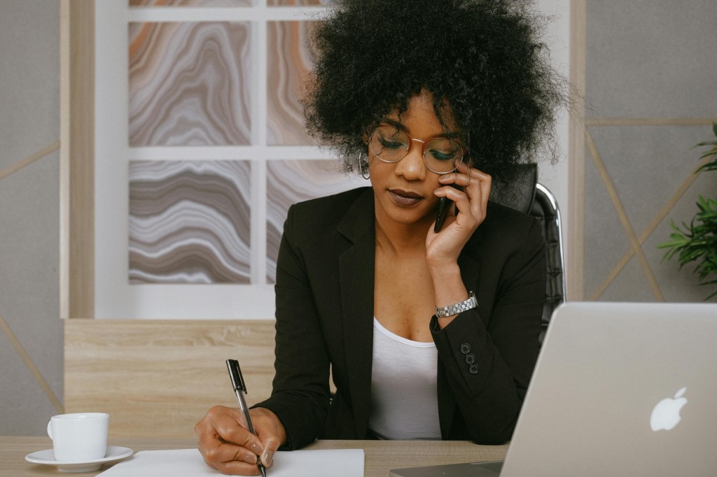 An African-American woman is seated at a desk with a laptop and a coffee mug. She is talking on the phone while writing.